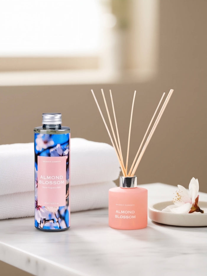 A reed diffuser bottle and matching diffuser sticks sit on a white marble surface beside folded white towels and a small dish with flower petals. The bottle has a blue floral label reading ALMOND BLOSSOM, and the diffuser has a soft pink bottle with reeds and a silver cap.