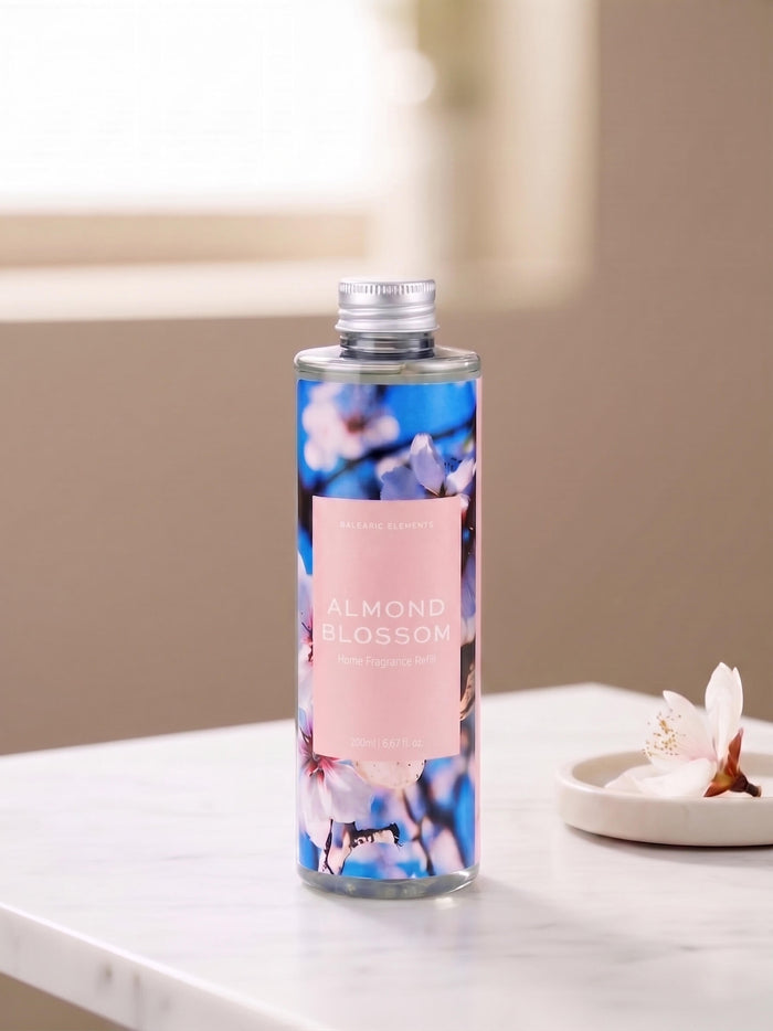 A bottle of Almond Blossom Reed Diffuser refill, featuring a pink label and blue floral design, set on a white marble countertop with a small dish of blossoms in a softly lit room.