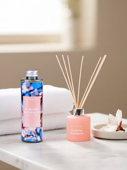 A reed diffuser bottle and matching diffuser sticks sit on a white marble surface beside folded white towels and a small dish with flower petals. The bottle has a blue floral label reading ALMOND BLOSSOM, and the diffuser has a soft pink bottle with reeds and a silver cap.