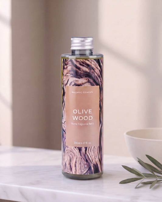 A bottle of Balancer Elements Olive Wood Home Fragrance Refill with a rustic wood grain label and silver cap, set on a marble countertop with a white bowl and olive leaves in a softly lit room.