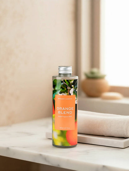 A clear bottle of reed diffuser refill labeled 'ORANGE BLEND' with a silver cap, standing on a white marble surface next to a folded cream towel. The background is a softly blurred warm beige interior with a small potted succulent.