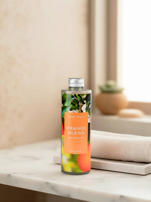 A clear bottle of reed diffuser refill labeled 'ORANGE BLEND' with a silver cap, standing on a white marble surface next to a folded cream towel. The background is a softly blurred warm beige interior with a small potted succulent.