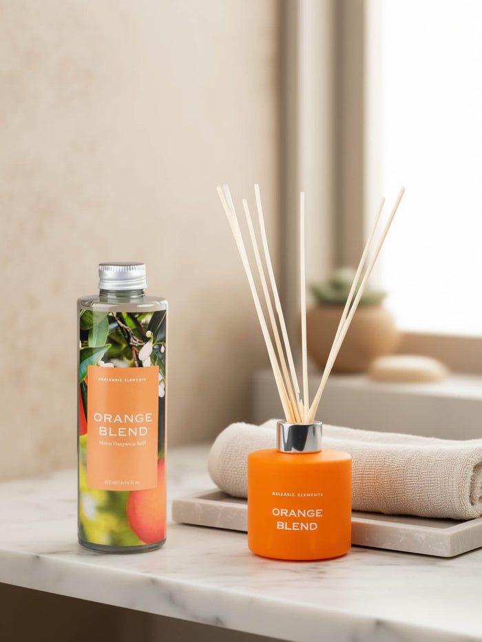 A reed diffuser set on a marble surface with a rolled towel and soft neutral background. The set includes a clear bottle with a silver cap and an orange reed diffuser base with natural reeds, both labeled 'ORANGE BLEND'.