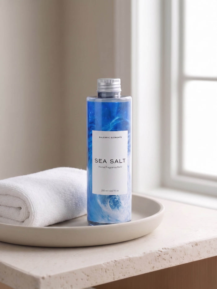 A bottle of Balearic Elements Sea Salt body wash stands on a beige tray next to a folded white towel, set on a windowsill with soft natural light.