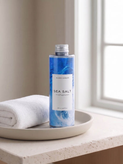 A bottle of Balearic Elements Sea Salt body wash stands on a beige tray next to a folded white towel, set on a windowsill with soft natural light.