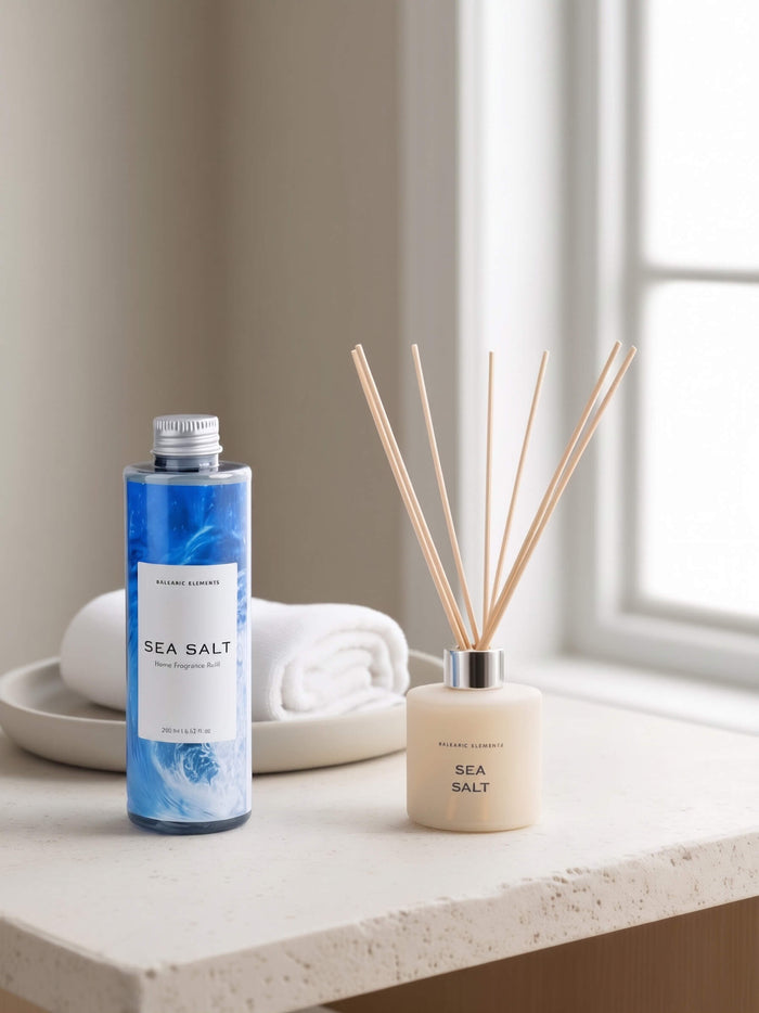 A bottle of Balearic Elements Sea Salt diffuser refill and a matching reed diffuser bottle with wooden reeds, styled on a light stone countertop with a folded white towel and dish in a softly lit room.