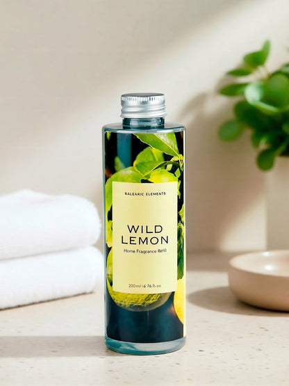 A bottle of Balearic Elements Wild Lemon home fragrance refill with a silver cap, standing on a light beige surface with a folded white towel, a small dish, and a green plant in the background.
