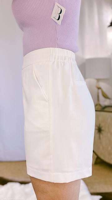 Side view of woman wearing tailored white shorts paired with a ribbed lavender top in a stylish indoor setting