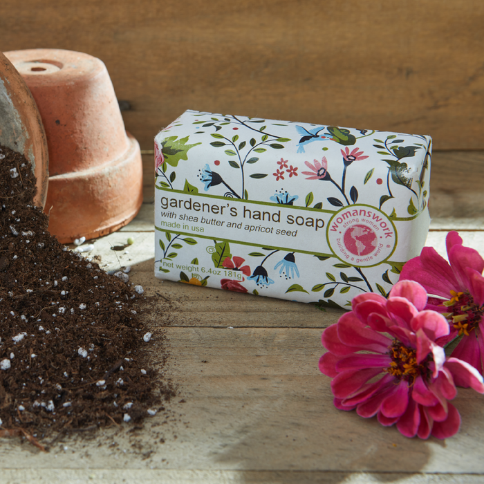 Bar of 'gardener's hand soap' with floral packaging on a wooden surface with soil and flowers.