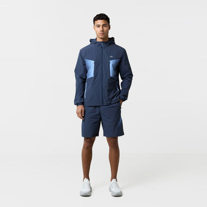 Model is wearing a tri-set with pair of navy shorts and a matching navy wind breaker featuring a logo and gradient shapes.