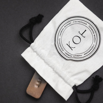 A white drawstring pouch with a black circular 'KOT' logo printed on the front, shown on a dark gray background with a tin of Fischersund KOT solid perfume partially tucked inside.