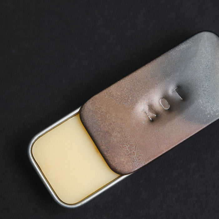 A small rectangular metal tin of Fischersund KOT solid perfume with a hinged lid, partially open. The lid is brushed metal with a textured bronze-brown center and the letters 'KOT' embossed on top.