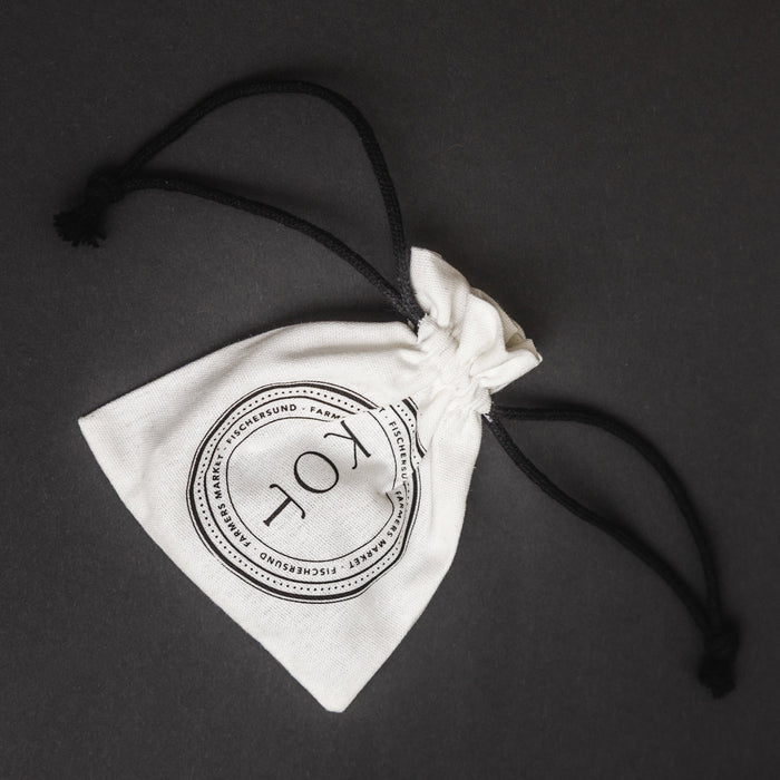 A white cotton drawstring pouch with a black printed circular logo reading 'KOT' and black drawstring cords, photographed on a dark gray background.