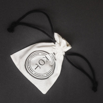 A white cotton drawstring pouch with a black printed circular logo reading 'KOT' and black drawstring cords, photographed on a dark gray background.