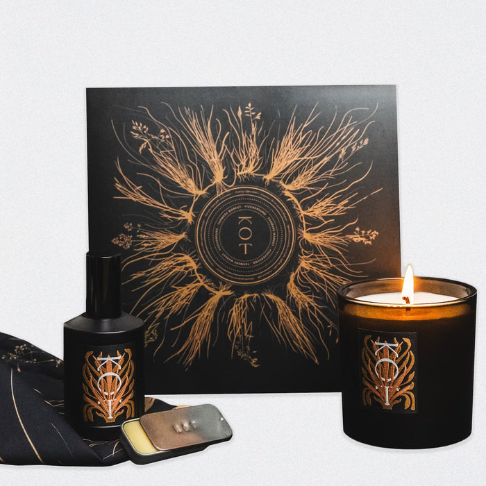 A group photo of the Fischersund KOT collection, a Farmers Market collaboration. The image features a black bottle of KOT perfume, a metallic tin of KOT solid perfume, a lit KOT candle in a black glass holder, a KOT vinyl album in a sleeve, and a black KOT bandana. The perfume, candle, and vinyl sleeve each feature the name KOT with a metallic printed design on the label.