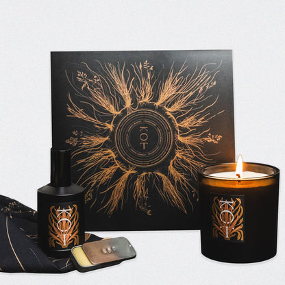A group photo of the Fischersund KOT collection, a Farmers Market collaboration. The image features a black bottle of KOT perfume, a metallic tin of KOT solid perfume, a lit KOT candle in a black glass holder, a KOT vinyl album in a sleeve, and a black KOT bandana. The perfume, candle, and vinyl sleeve each feature the name KOT with a metallic printed design on the label.
