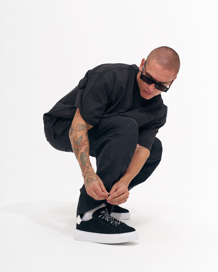 Martin Valen men's black baggy pant, crouching three-quarter view as model ties shoe, tattooed forearm visible