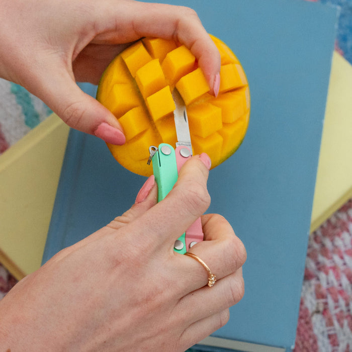 Hands using a Leatherman Micra® in Bayside color to cut a mango on top of a stack of books.