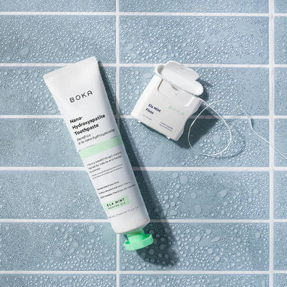 Boka toothpaste tube and dental floss on a tiled surface with water droplets.