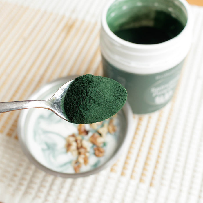 Spoonful of green spirulina powder over a bowl with open container in the background.