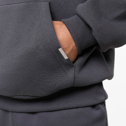 Model's hand in the kangaroo pocket of the dark grey hoodie, showing the branded pocket detail