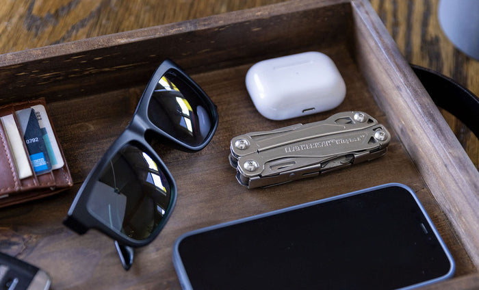 Stainless steel Leatherman Wingman® multi-tool beside sunglasses, smartphone, and AirPod case in a wooden tray.
